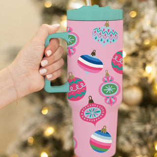 A 40 oz stainless steel tumbler with a pink background and multicolored Christmas ornament designs. The tumbler is held in a person's hand with a Christmas tree in the background.