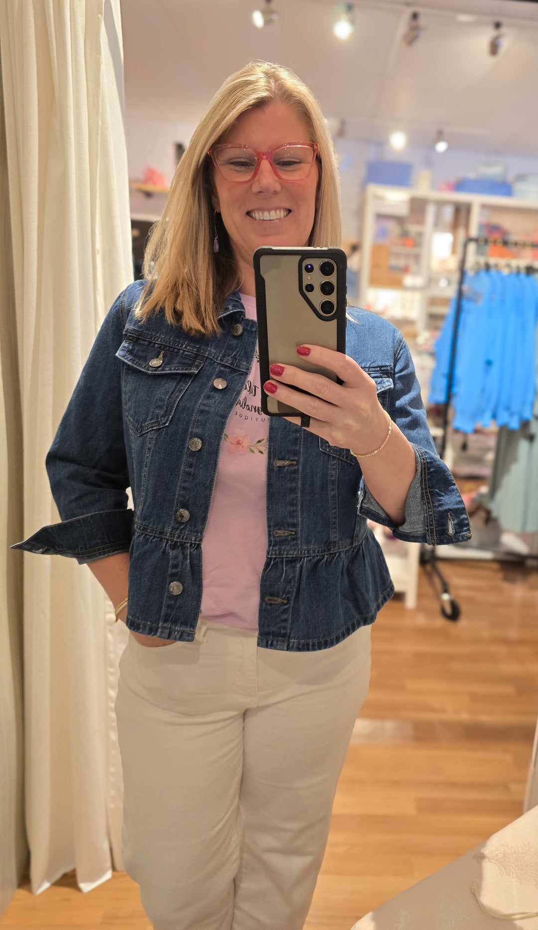 Woman taking a mirror selfie wearing a denim jacket and beige pants in a store.