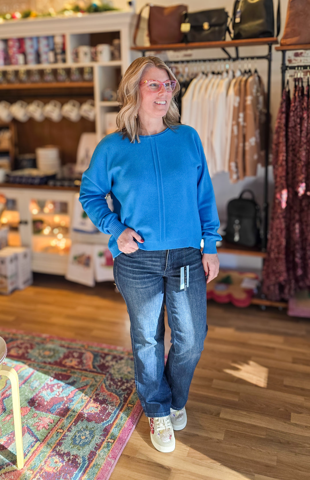 A woman wearing a bright blue crewneck sweatshirt and blue jeans stands inside a retail store, with clothing racks, shelves, and a patterned rug visible in the background.