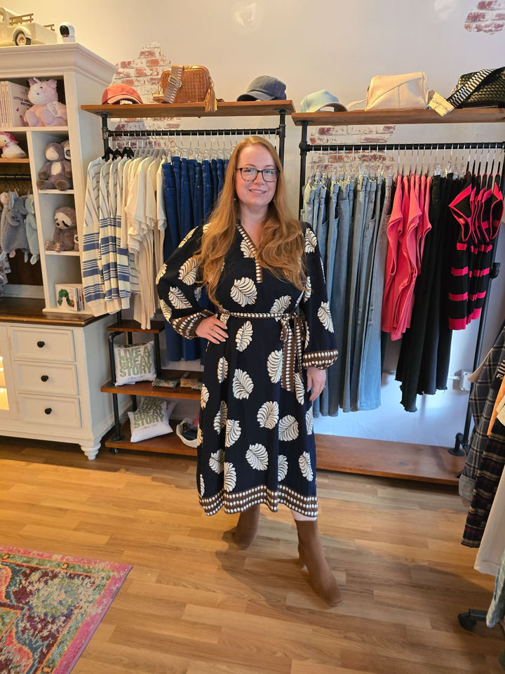 A woman wearing a navy blue and white patterned long-sleeve dress stands inside a boutique clothing store with racks of garments and wooden floors.