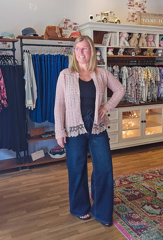 A woman modeling a light pink crochet cardigan with lace trim over a black top and wide-leg blue jeans, standing inside a boutique clothing store.