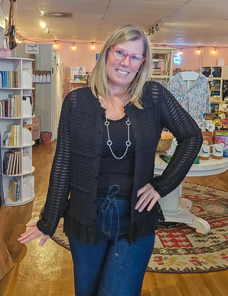 A woman with blonde hair and glasses wearing a black crochet cardigan over a black top and blue jeans, standing inside a retail boutique.