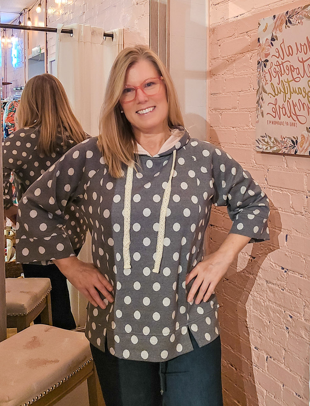 A woman wearing a gray pullover hoodie with white polka dots and cream drawstrings, standing in a boutique fitting room with a pink brick wall and mirror behind her.