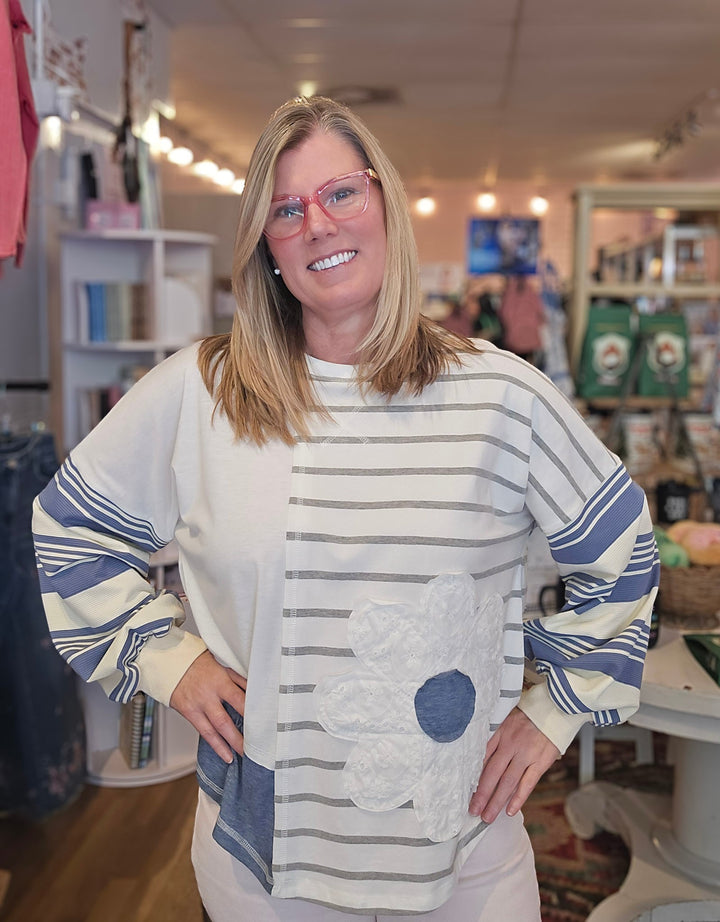 A smiling blonde woman wearing pink-framed glasses and a cream-colored long-sleeve sweatshirt with blue stripes and a large applique flower, standing inside a retail store.