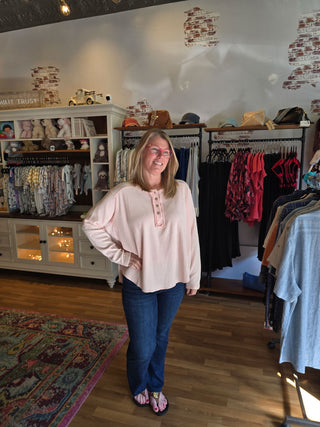 A woman wearing a light pink long-sleeve blouse with button details and dark blue jeans, standing inside a clothing boutique with racks of garments, shelves, and a patterned rug visible.