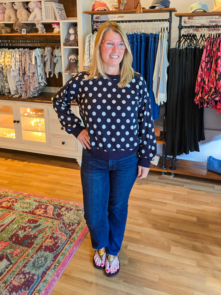 A woman modeling a navy blue long-sleeve sweatshirt with white polka dots, paired with dark blue jeans and pink sandals, standing inside a clothing boutique.