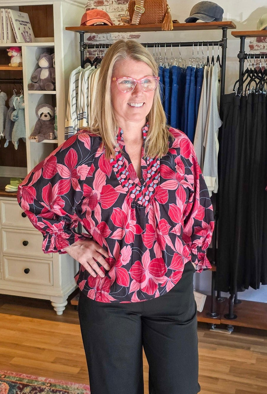 A woman wearing a pink and black floral long-sleeve blouse and black pants stands inside a clothing boutique. The background shows racks of clothes, a display cabinet, and a wooden floor.