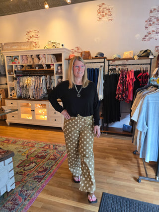 A woman stands in a boutique wearing beige wide-leg pants with white polka dots, a black long-sleeve top, and pink sandals. The shop has wood floors, clothing racks, and a display cabinet in the background.