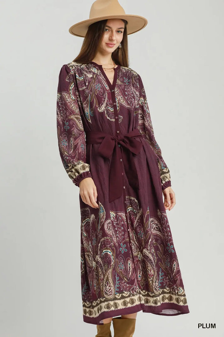 Woman wearing a long, patterned dress with a belt and a hat on a plain background.