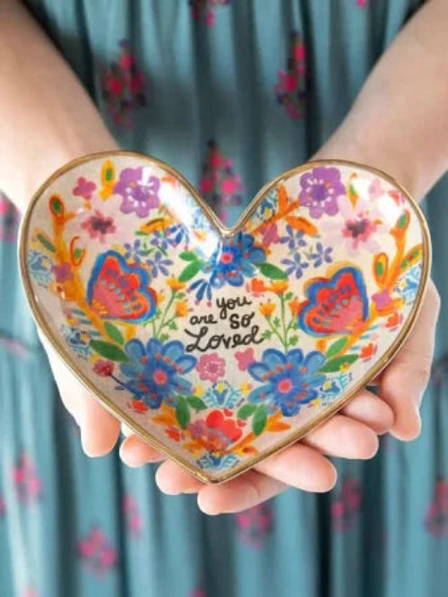 Heart-shaped decorative plate with floral design and 'you are so loved' text held by hands.