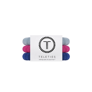 Three rolled hair ties in light blue, magenta pink, and navy blue, bundled together with a white paper band featuring the black TELETIES logo and brand name.