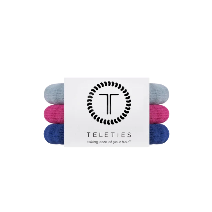 Three rolled hair ties in light blue, magenta pink, and navy blue, bundled together with a white paper band featuring the black TELETIES logo and brand name.