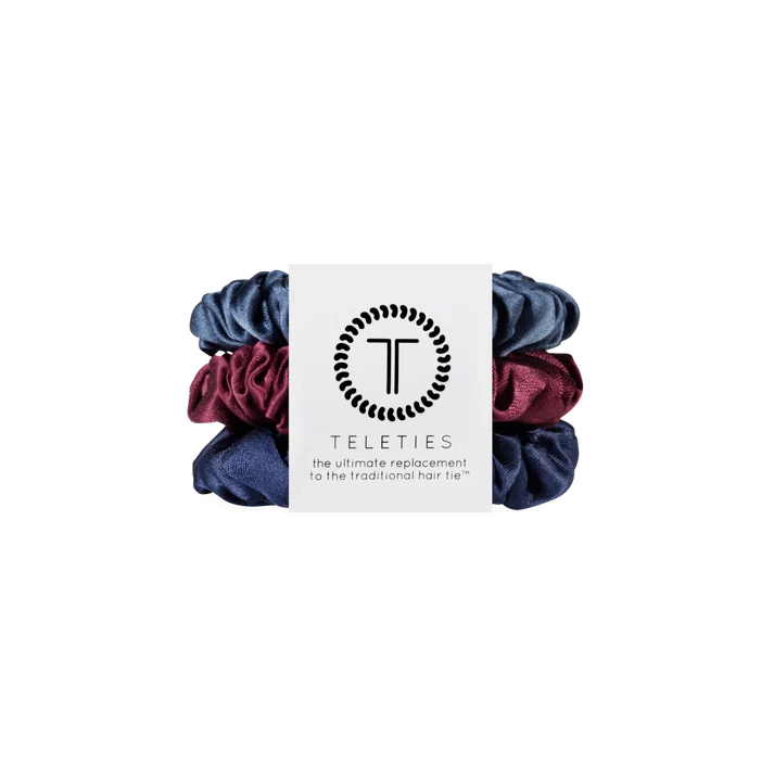 Three satin hair scrunchies in navy blue, burgundy, and dark purple, wrapped with a white paper band featuring the Teleties brand logo.
