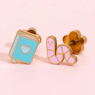 A pair of gold-tone enamel stud earrings on a soft pink background. One earring is shaped like a light blue book with a gold outline and a small heart detail.