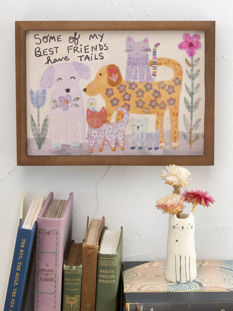 Framed children's illustration of animals and flowers on a wall with books and a vase below.