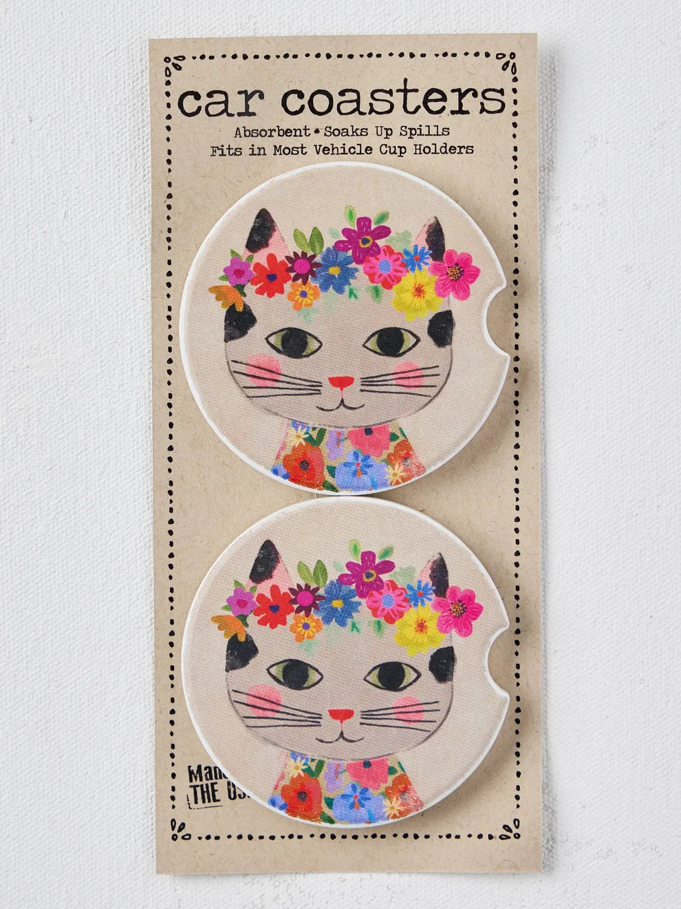 A package of two round car coasters featuring a cute cartoon cat face with black ears, pink nose, and a colorful floral crown and floral collar, mounted on a beige cardboard backing labeled &