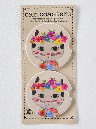 A package of two round car coasters featuring a cute cartoon cat face with black ears, pink nose, and a colorful floral crown and floral collar, mounted on a beige cardboard backing labeled 'car coasters'.