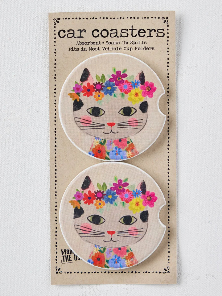 A package of two round car coasters featuring a cute cartoon cat face with black ears, pink nose, and a colorful floral crown and floral collar, mounted on a beige cardboard backing labeled &