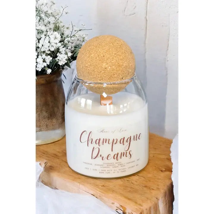 Candle labeled 'Champagne Dreams' with a cork lid on a wooden surface.