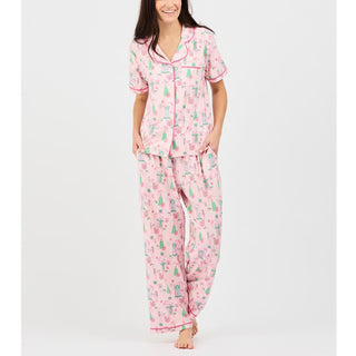 woman wearing short sleeve top and pants pajama set in light pink. Features green and pink christmas pattern with trees, wreaths, garlands, and bows