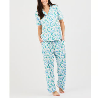 woman wearing short sleeve top and pants pajama set with green and blue christmas icon designs. Featuring christmas trees, wreaths, bows, and gifts