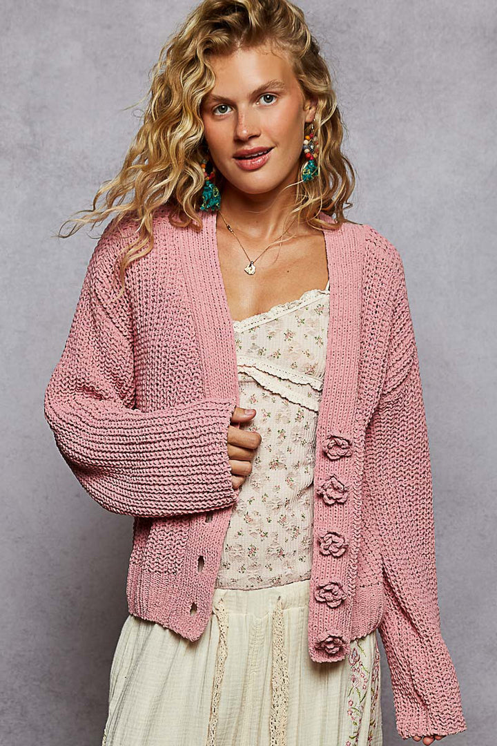 Woman wearing a pink knitted cardigan over a white lace top against a gray background