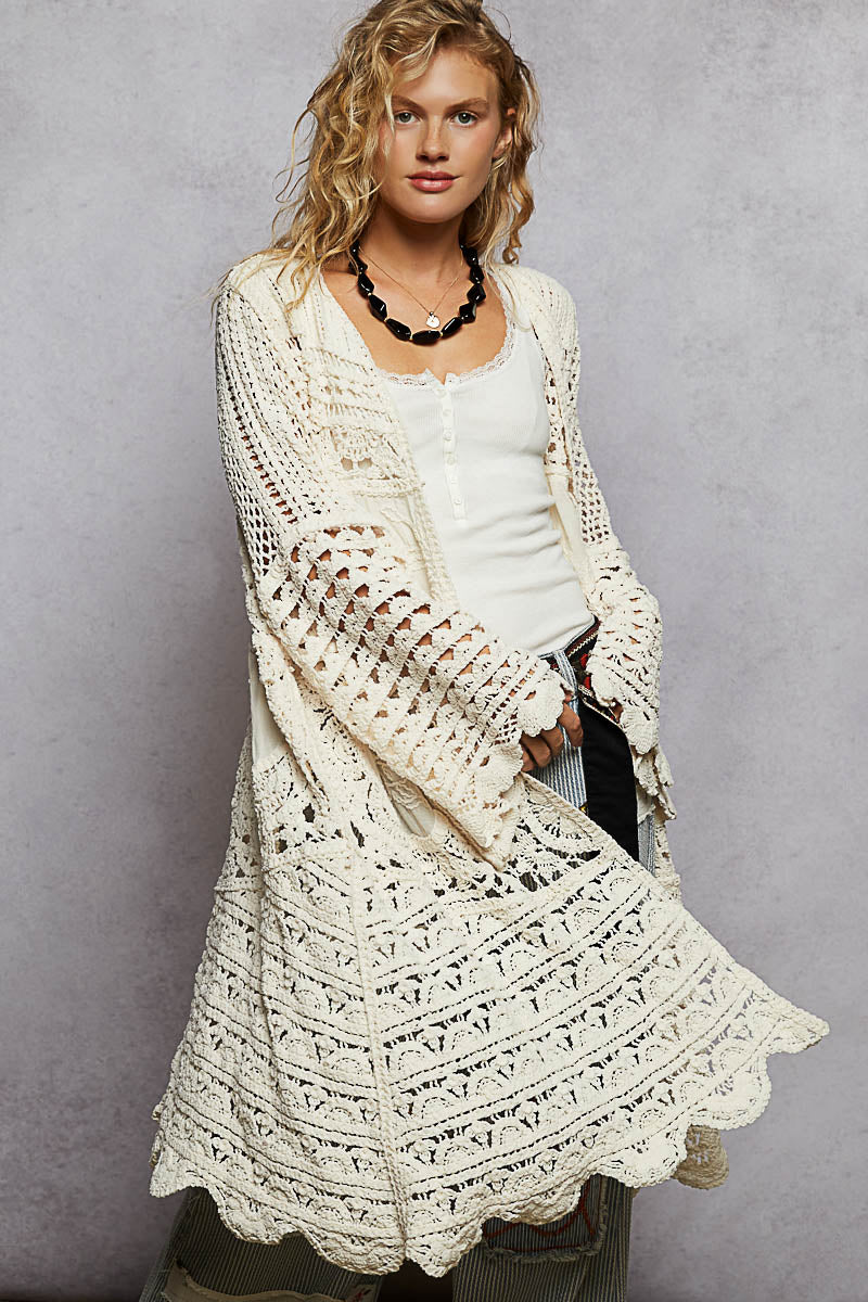 Woman wearing a long, white crochet cardigan over a white top against a gray background