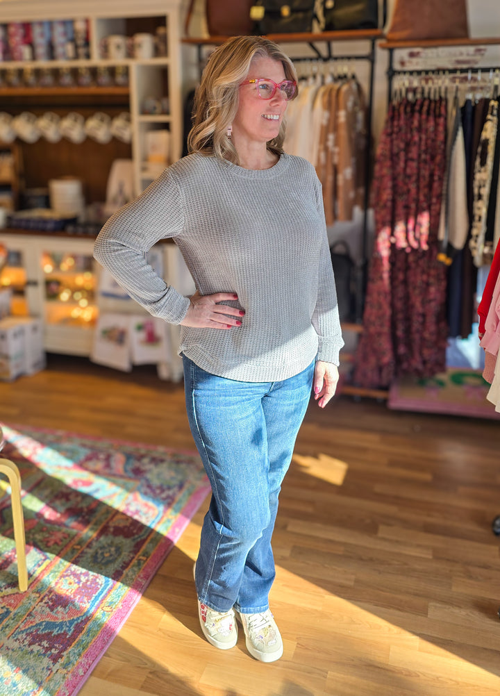 Woman wearing a gray sweater and blue jeans standing in a store with racks of clothes in the background.