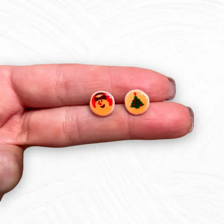 Two small round earrings with Christmas designs held between fingers on a white background
