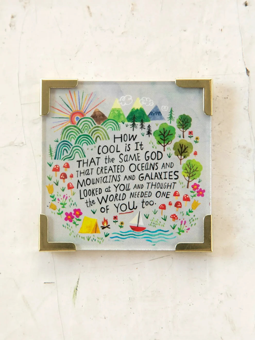 A square framed art print with a gold-toned metal frame, featuring a whimsical hand-drawn landscape with mountains, trees, flowers, a sun, and a quote about being seen by the recipient, displayed on a light textured background.