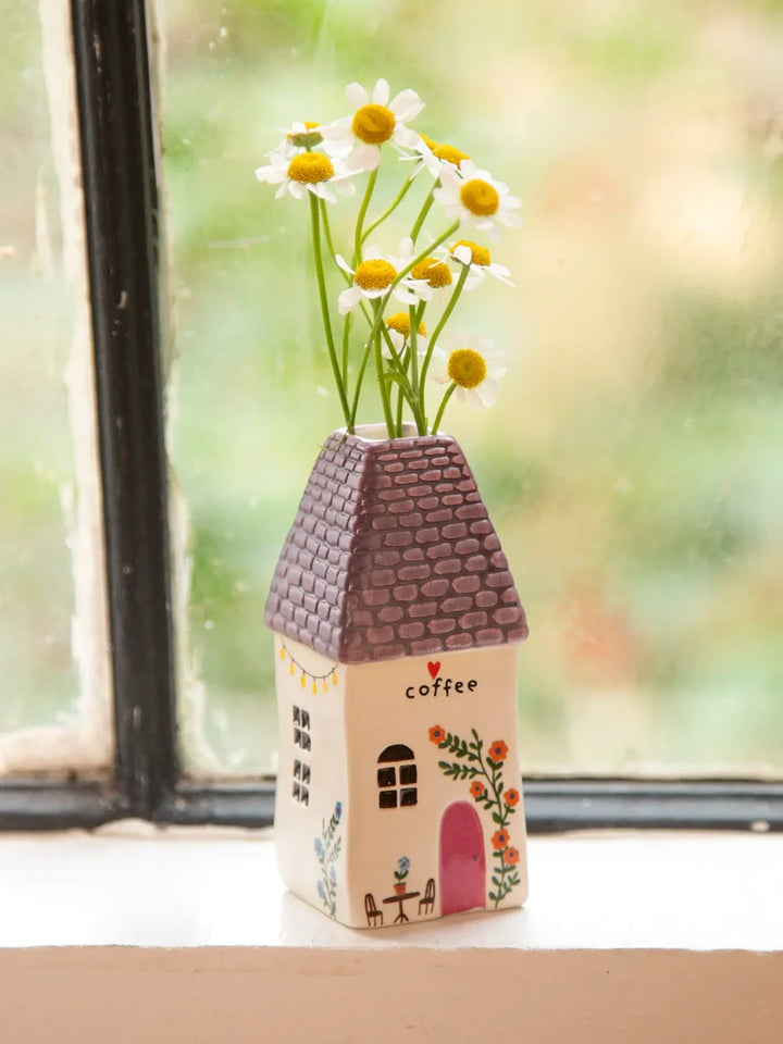 A small ceramic house-shaped vase with a purple roof and floral details, holding white daisies with yellow centers, placed on a windowsill with a blurred outdoor background.