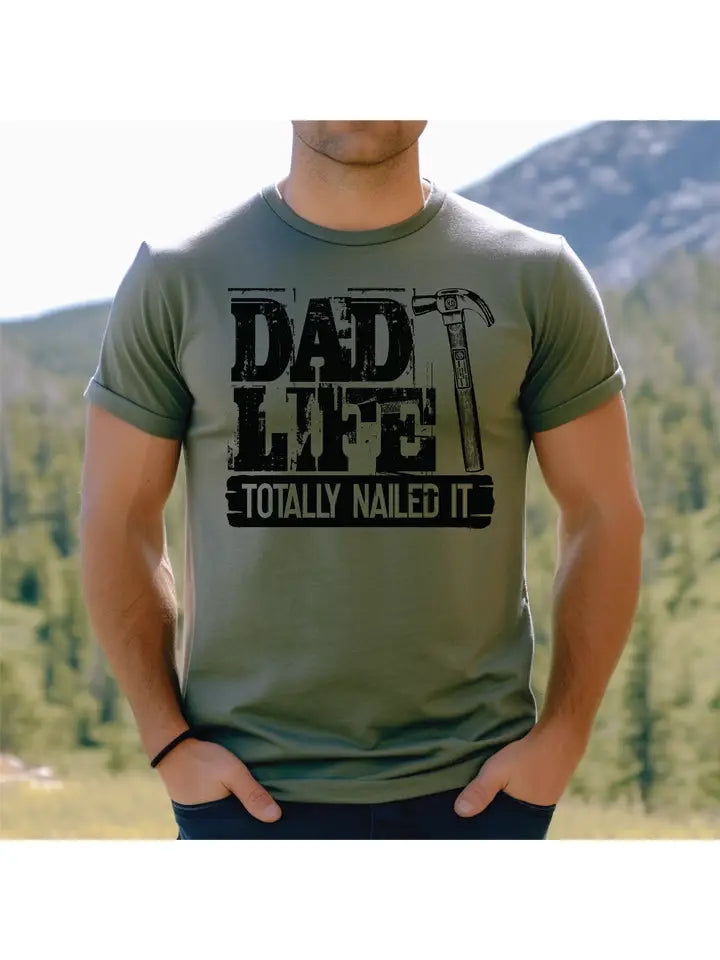 Person wearing a green t-shirt with 'Dad Life Totally Nailed It' text in a mountainous landscape.