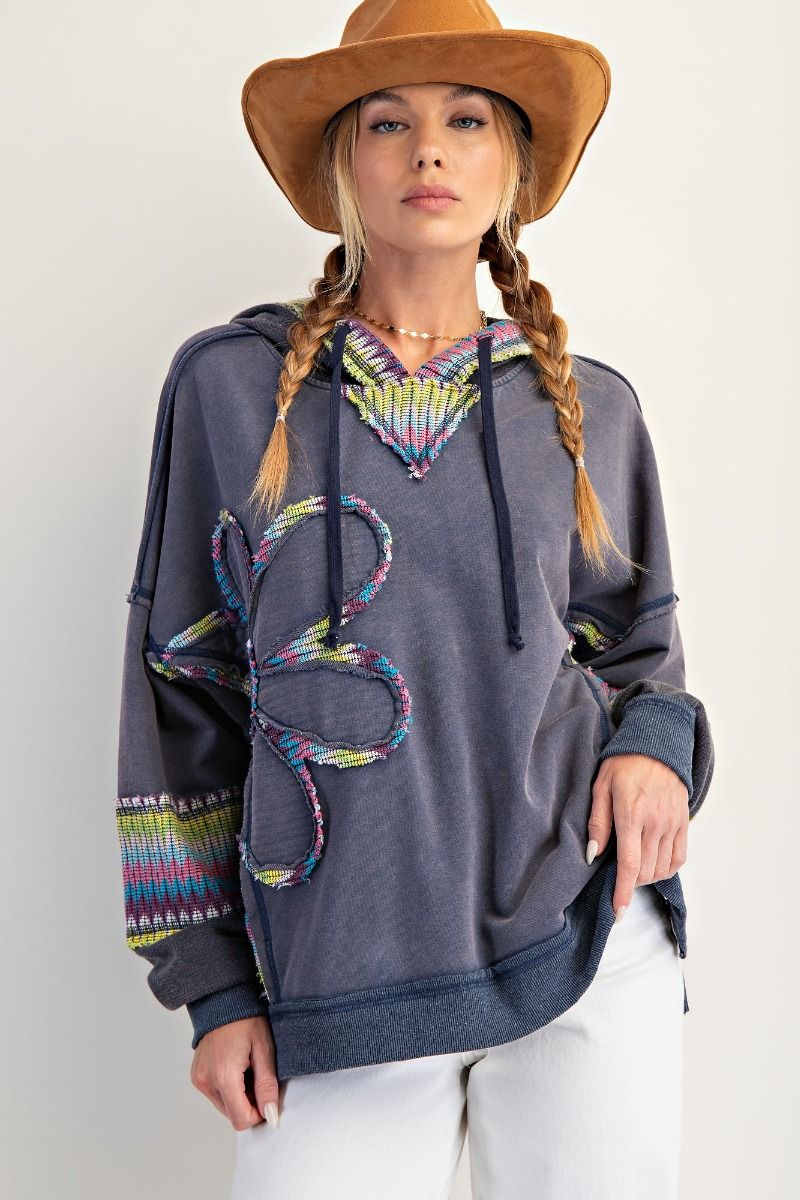 Woman wearing a gray hoodie with colorful designs and a brown hat on a plain background