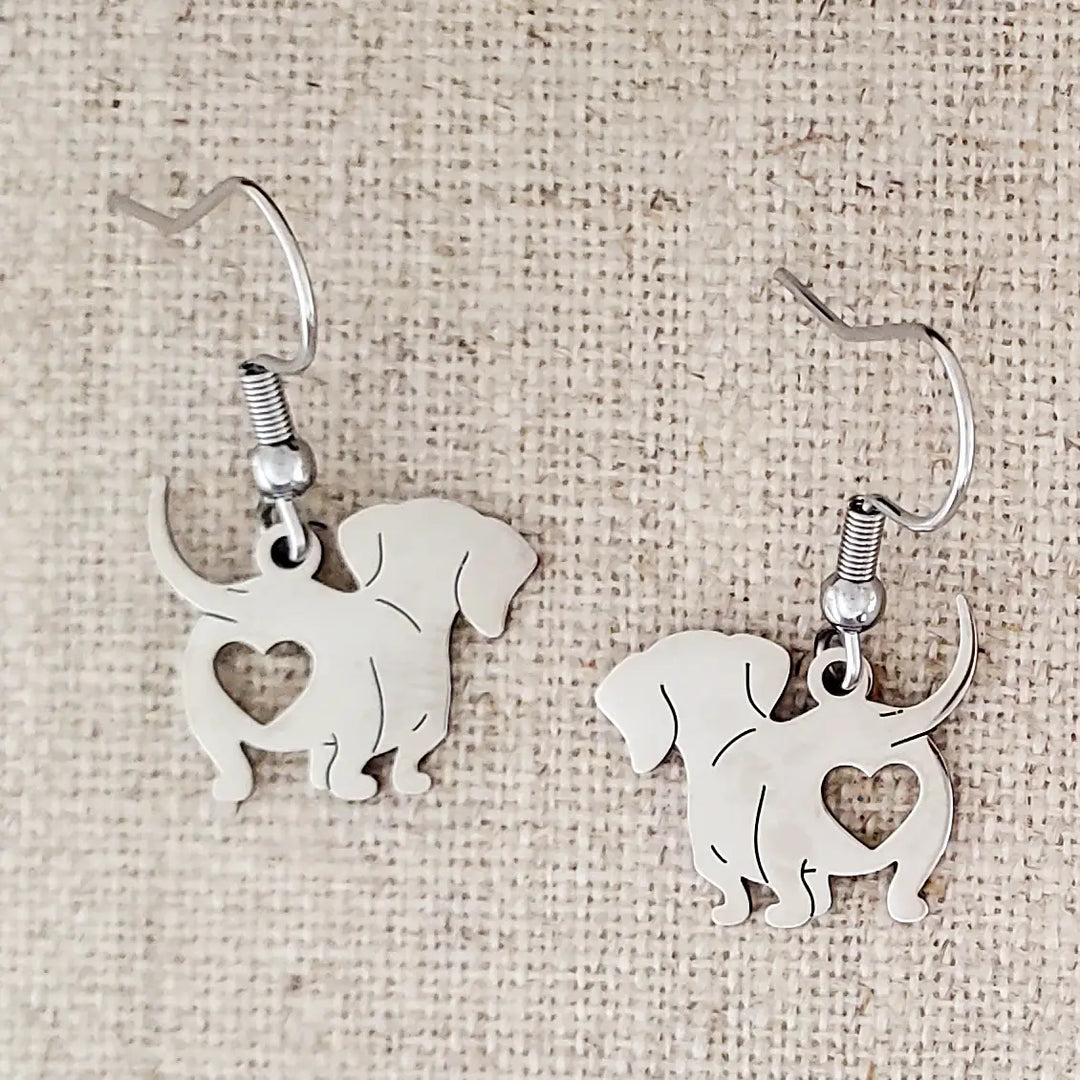 Silver dog-shaped earrings with heart cutouts on a beige fabric background