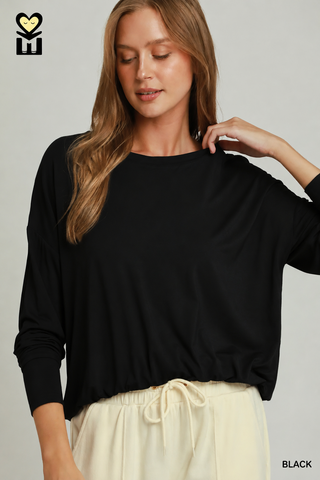 A woman wearing a black long-sleeve crewneck top and cream drawstring pants, shown against a light gray studio background. A small black-and-white logo appears in the top-left corner, and the word 'BLACK' is printed in the bottom-right corner.