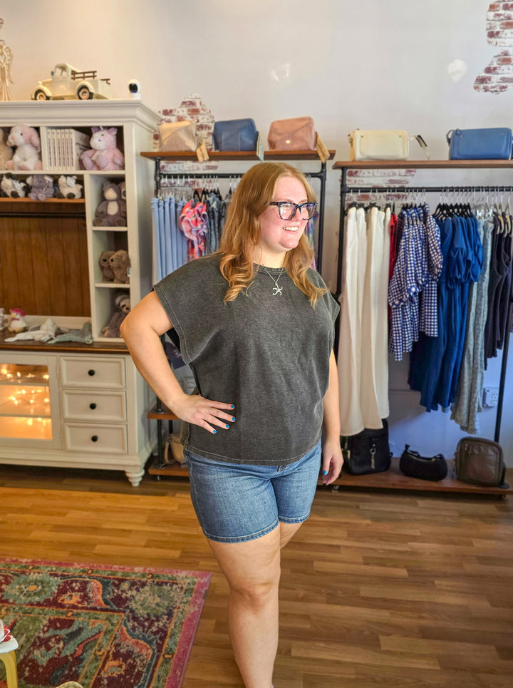 Woman standing in a clothing store wearing a gray sleeveless top and blue shorts.
