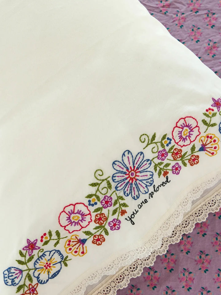 An off-white embroidered pillowcase featuring a colorful floral border and the embroidered phrase &