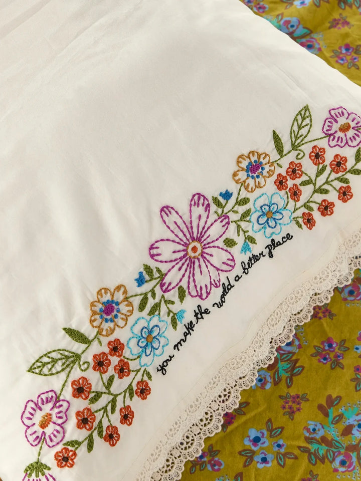 A close-up of a white embroidered fabric, likely a pillowcase or decorative textile, featuring colorful floral embroidery and a black script quote. The fabric is laid over a yellow floral patterned surface.