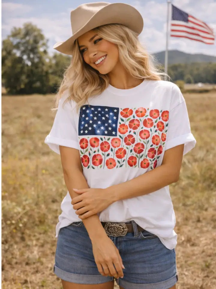Woman wearing a white t-shirt with a floral American flag design, standing in a field with an American flag in the background.