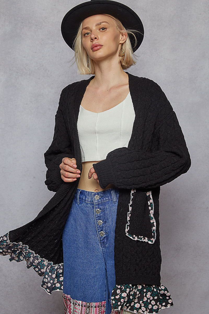 Woman wearing a black cardigan with floral trim, white top, and blue jeans against a gray background
