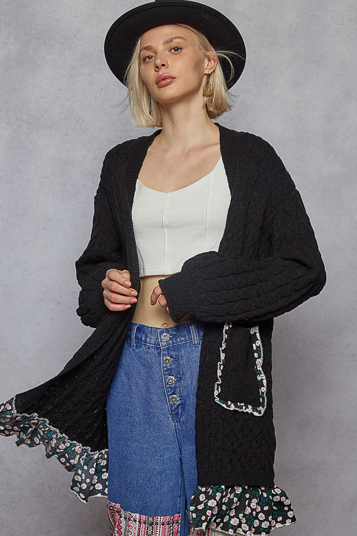 Woman wearing a black cardigan with floral trim, white top, and blue jeans against a gray background