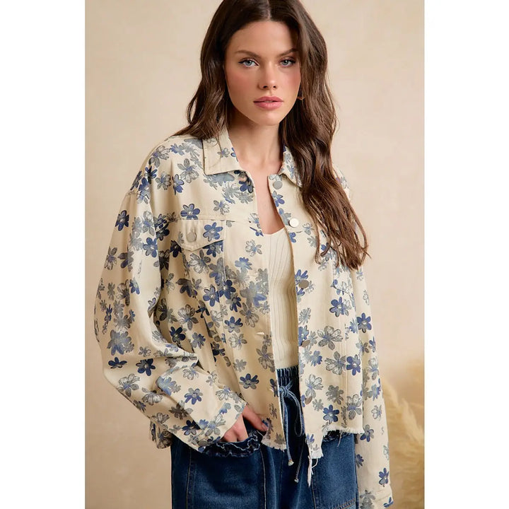 A woman wearing a cream-colored, long-sleeve button-up shirt jacket with a blue floral pattern, layered over a ribbed cream top and paired with dark blue jeans.