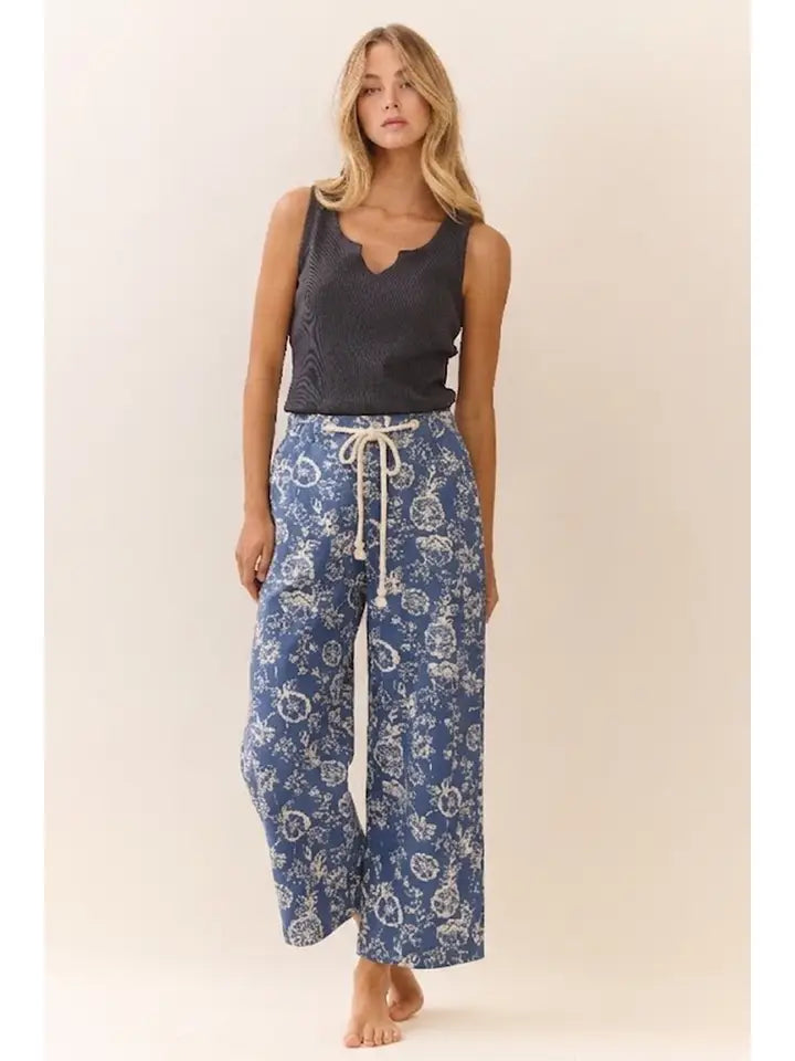A woman stands barefoot against a plain beige studio background wearing a dark charcoal sleeveless top and loose blue pants with a white paisley pattern and white drawstring.