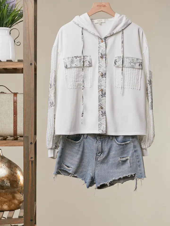 A light gray hooded button-up shirt with floral patterned chest pockets and distressed sleeves, styled with light-wash denim shorts on a hanger against a beige wall.