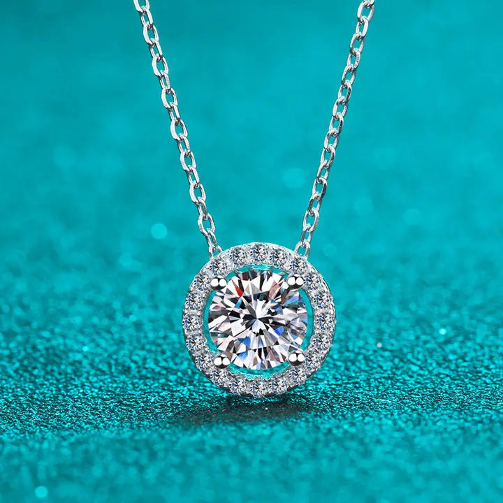 A round necklace featuring a halo-style moissanite gemstone in a silver setting, displayed against a teal background.