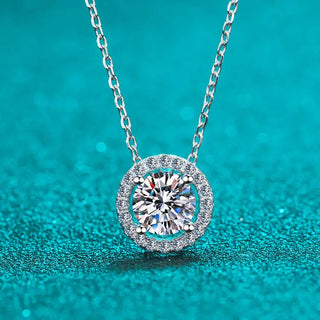 A round necklace featuring a halo-style moissanite gemstone in a silver setting, displayed against a teal background.