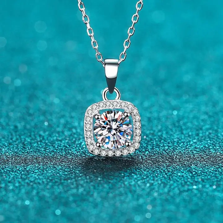 A silver-toned square halo pendant necklace with a central round clear gemstone and small surrounding stones, hanging on a chain against a sparkling turquoise background.