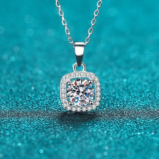A silver-toned square halo pendant necklace with a central round clear gemstone and small surrounding stones, hanging on a chain against a sparkling turquoise background.
