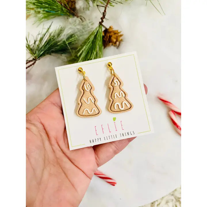 Gold tree-shaped earrings on a card held by a hand with Christmas decorations in the background