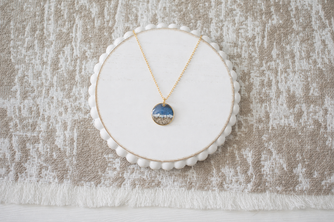 A delicate gold necklace with a small oval pendant featuring a scenic landscape of blue water and white sand, displayed on a white circular bead-edged stand over a textured beige and white fabric background.